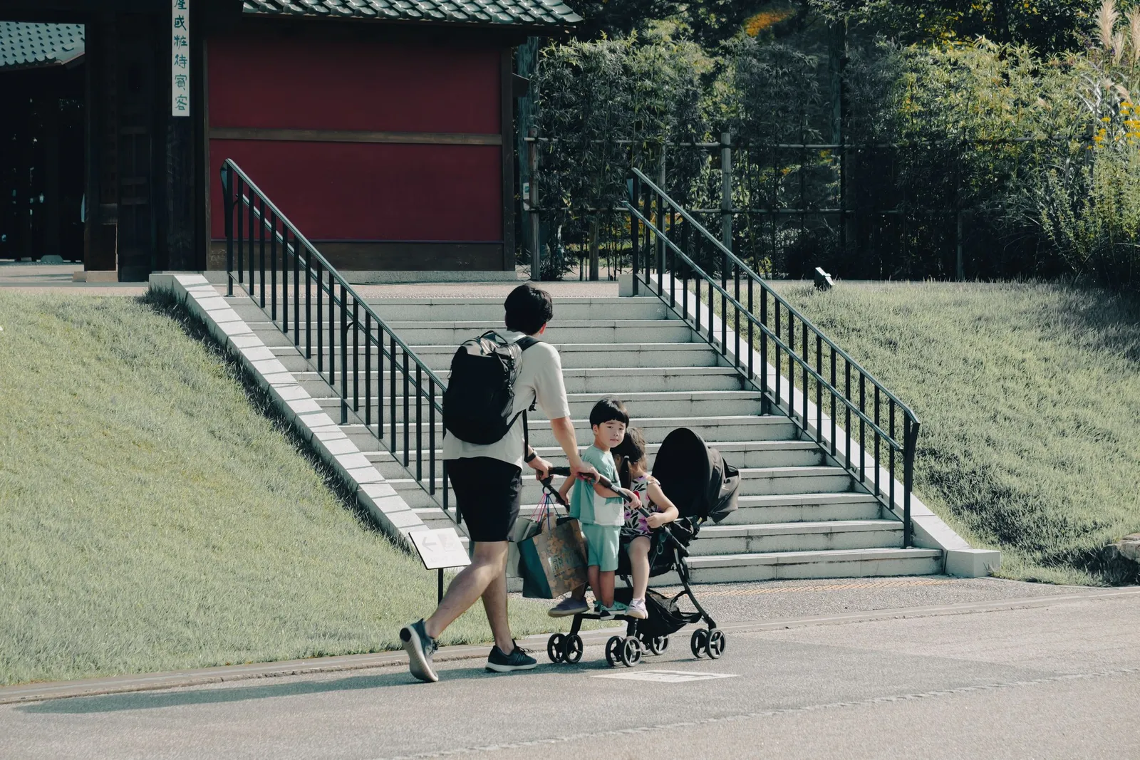Father pushing a stroller with a child past a stone staircase on a sunny day in Nagoya, Japan
