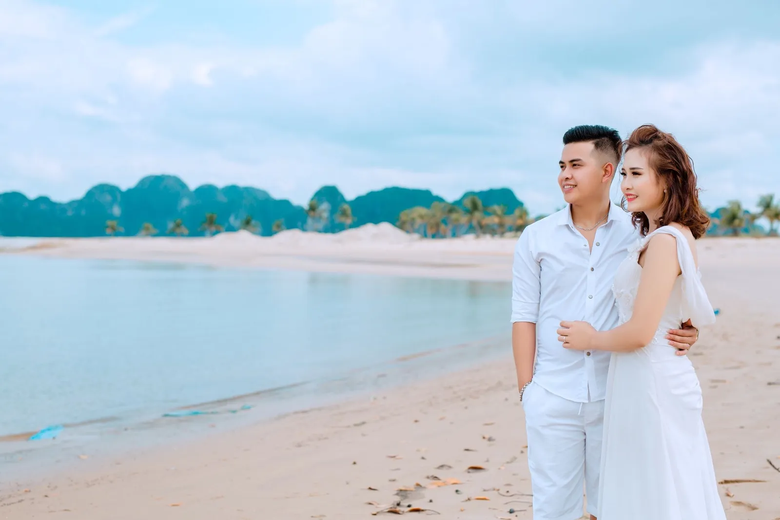 Couple dressed in white embracing on a quiet sandy beach at sunrise with limestone islets on the horizon
