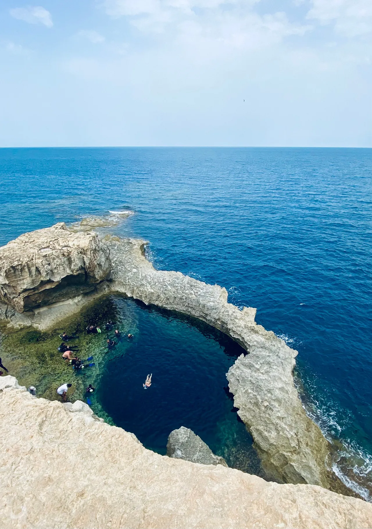 Natural blue hole in the Omani coastline with divers and swimmers in crystal-clear water opening to the Arabian Sea