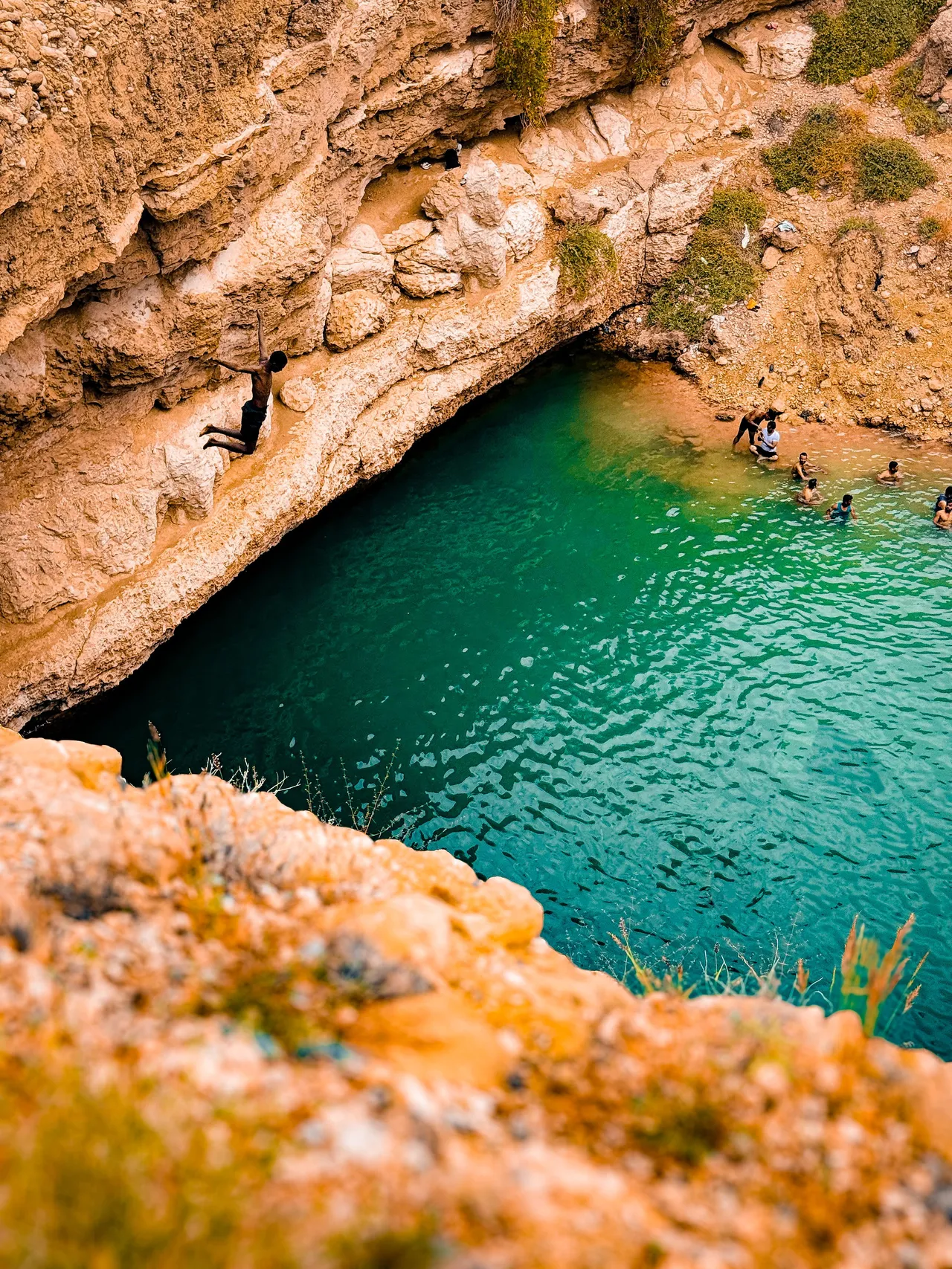 Turquoise rock pool inside a limestone wadi in Oman with cliff jumpers and swimmers below an overhanging cave